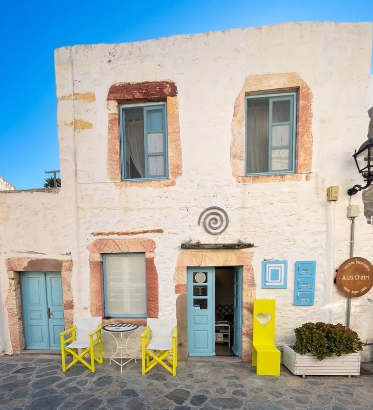 The studio in Skala: a limewashed house with blue doors, yellow chairs and an iron spiral above the entrance